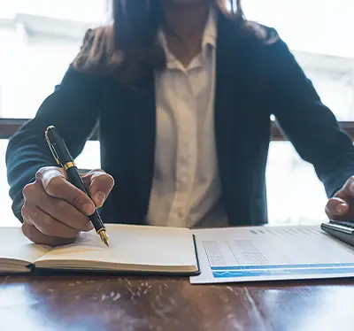 home 2nd banner img A business professional in a blazer writing in a notebook, with a document and a pen on a wooden table in a well-lit setting.