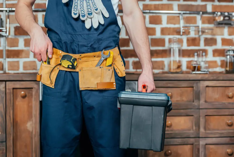 man with tool A man in blue overalls holds a toolbox in one hand and tools in a leather belt, standing in a workshop with a brick wall backdrop.